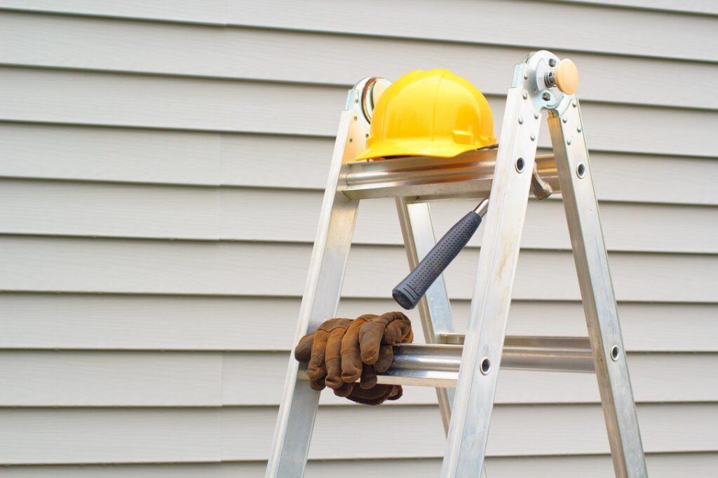Contractor's gear on ladder next to siding to represent the importance of professional installation Stepladder, hardhat, gloves and hammer with house siding background